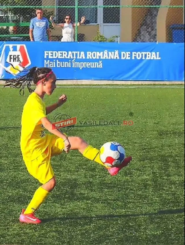 O fată îmbrăcată în echipament sportiv galben lovește un fotbal alb cu pete albastre și roșii pe un teren de fotbal verde.