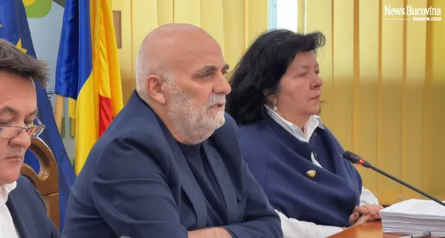 Un bărbat cu barbă și păr scurt, îmbrăcat în bluza albastră, stă lângă o femeie cu păr negru și sacou albastru în fața unor steaguri.