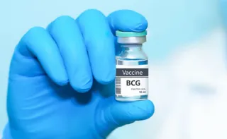 O mână îmbrăcată în mănușă albastră ține un flacon de sticlă cu etichetă albă pe care scrie BCG.