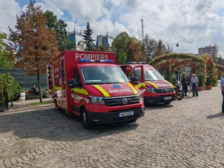 Două ambulanțe roșii și galbene cu inscripții sunt parcate pe un drum pavat lângă copaci și vegetație sub un cer înnorat.