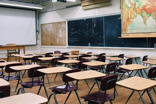 O sală de clasă goală, cu bănci și scaune aliniate, o tablă neagră cu formule și o hartă pe perete.