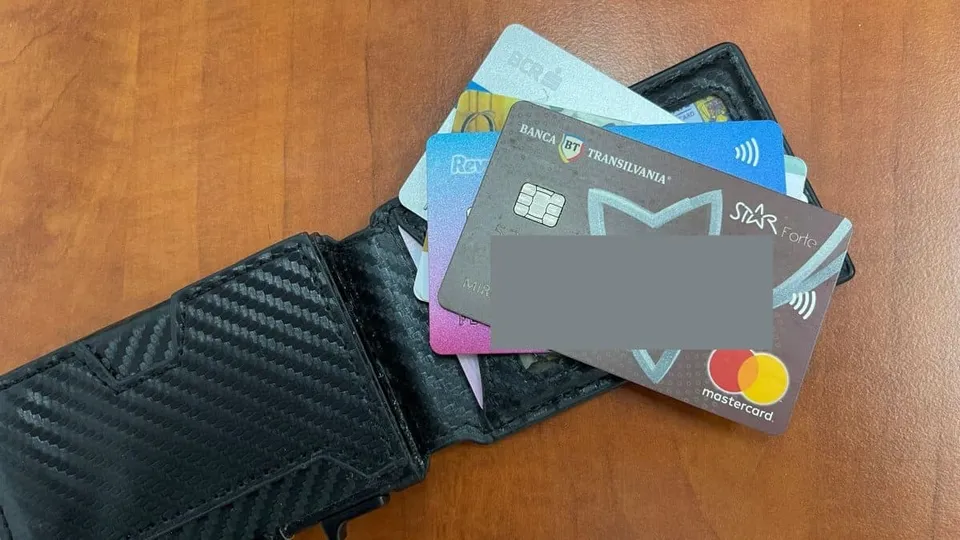 Un portofel negru deschis conține mai multe carduri bancare colorate, printre care unul de culoare gri cu logo-ul Mastercard este vizibil în partea de jos.