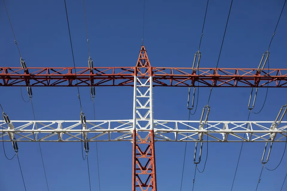 O structură metalică de înaltă tensiune, de culoare albă și portocalie, susține mai multe cabluri electrice sub un cer albastru.