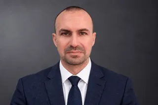 Un bărbat cu părul scurt și barbă scurtă poartă sacou albastru, cămașă albă și cravată închisă la culoare pe un fundal gri neutru.