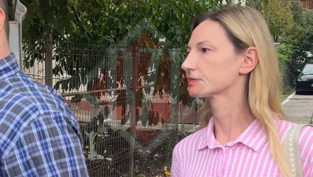 O femeie cu părul blond și o cămașă roz cu dungi albe stă în profil lângă un bărbat îmbrăcat într-o cămașă în carouri albastră și albă.