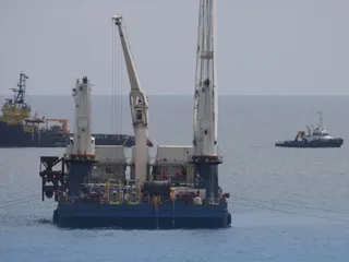 O platformă de lucru cu două macarale mari navighează pe apă, având alte două nave în depărtare sub un cer senin.
