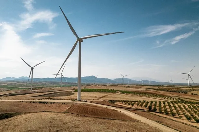 Mai multe turbine eoliene albe sunt amplasate pe un peisaj rural cu câmpuri arate și dealuri sub un cer înnoptat cu nori dispersi.