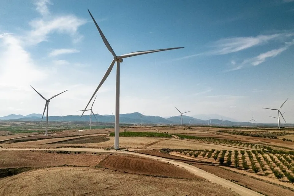 Mai multe turbine eoliene albe sunt amplasate pe un peisaj rural cu câmpuri arate și dealuri sub un cer înnoptat cu nori dispersi.