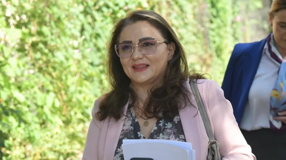 O femeie cu ochelari, purtând o bluză cu model floral și un sacou roz, ține în mână niște hârtii albe.