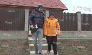 Un bărbat cu o geantă neagră și o femeie cu o jachetă portocalie coboară trepte în fața unei case cu acoperiș roșu.