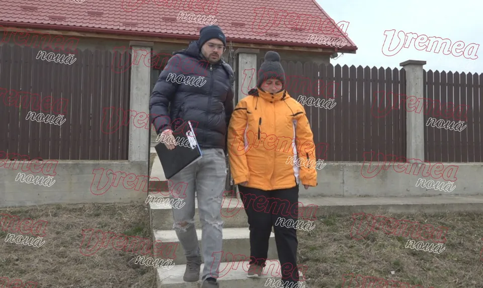 Un bărbat cu o geantă neagră și o femeie cu o jachetă portocalie coboară trepte în fața unei case cu acoperiș roșu.