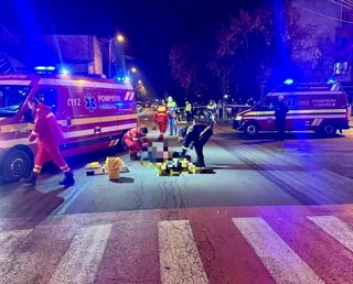 Imagine de noapte cu o ambulanță, pompieri și personal medical în jurul unei persoane întinse pe asfalt, la o trecere de pietoni.