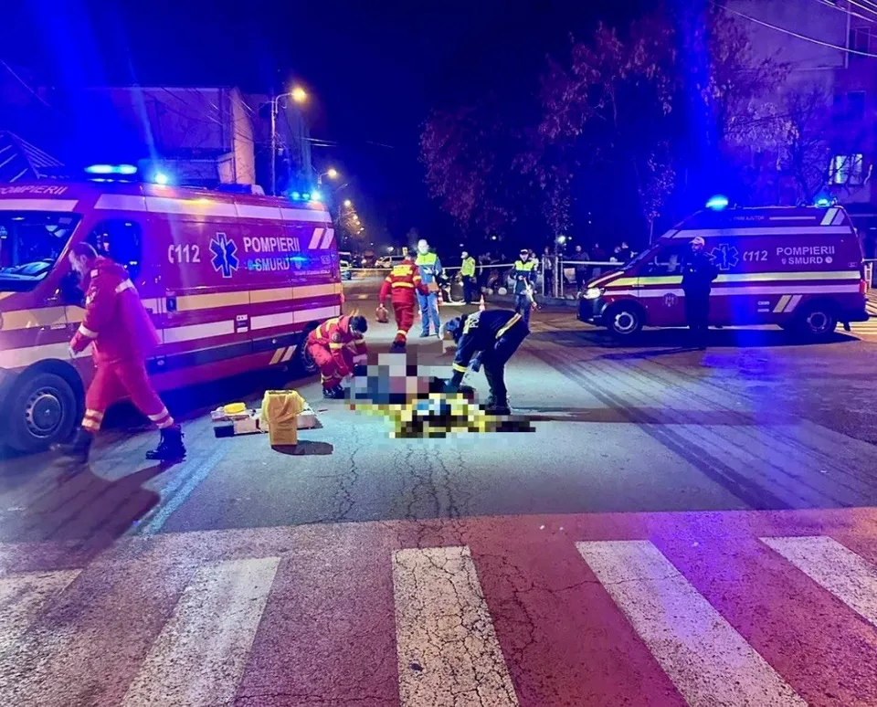 Imagine de noapte cu o ambulanță, pompieri și personal medical în jurul unei persoane întinse pe asfalt, la o trecere de pietoni.