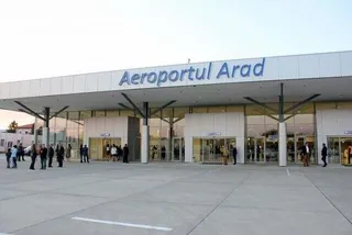 O clădire de terminal cu inscripția Aeroportul Arad, având suprafețe vitrate și persoane stând pe o platformă de beton în fața intrării.
