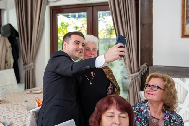 Un bărbat într-un costum negru ridică un telefon mobil pentru a face o fotografie cu două femei și o altă femeie în prim-plan.