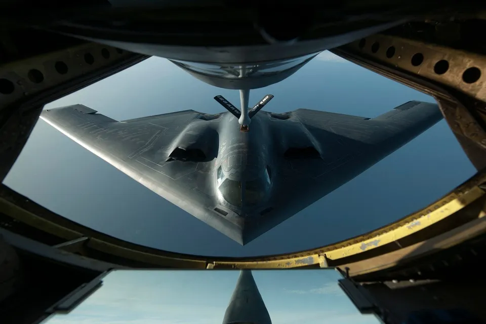 Un avion de forma triunghiulară zboară pe cer, fiind surprins prin cadrul deschis al unei alte aeronave.