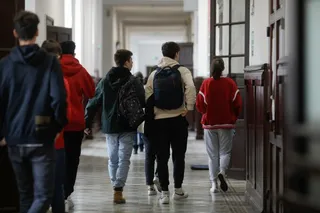 Un grup de elevi cu rucsacuri merge pe un coridor într-o clădire cu uși de lemn și pereți albi.