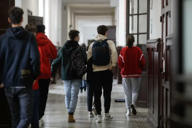 Un grup de elevi cu rucsacuri merge pe un coridor într-o clădire cu uși de lemn și pereți albi.