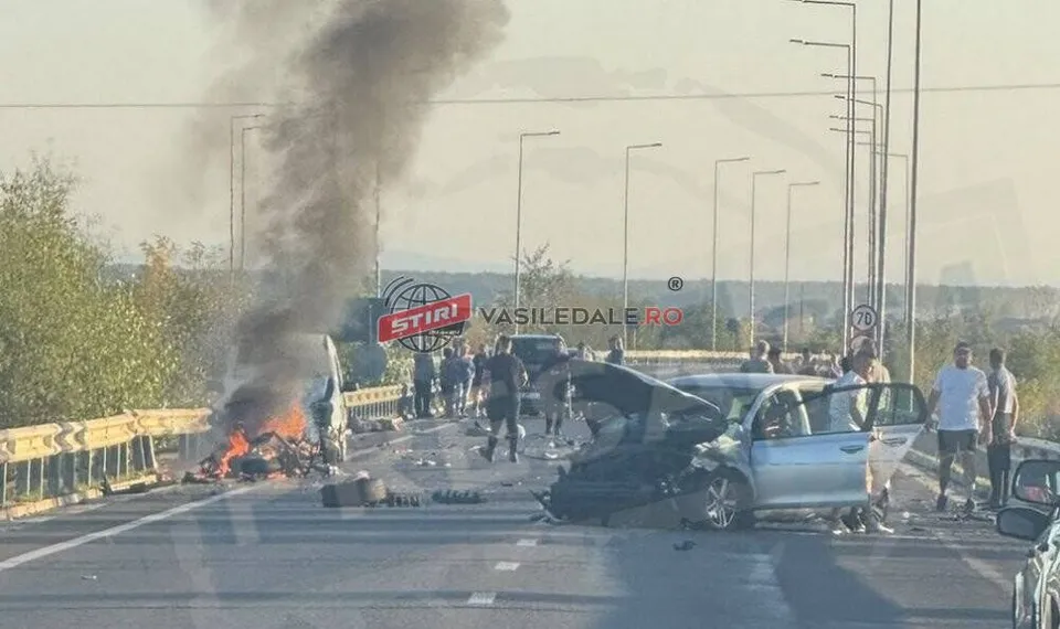 Un vehicul de culoare închisă arde pe marginea drumului, emanând un nor gros de fum negru, lângă un autoturism argintiu cu portiera deschisă.