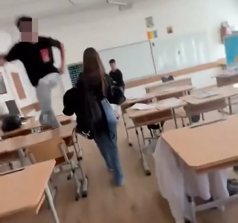 Un grup de elevi se află într-o sală de clasă cu bănci de lemn și un panou alb dispuse în interiorul încăperii.