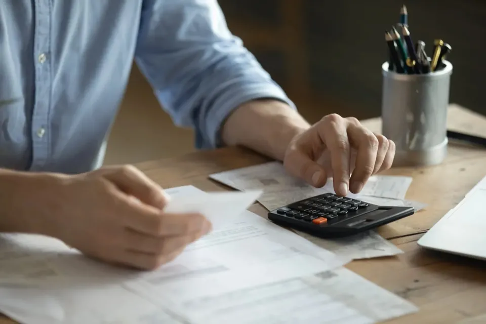 O persoană în costum deschis folosește o calculatoră pe birou, având în jur documente și un stilou cu creioane.