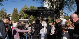 Un grup de oameni stă adunat în fața unei structuri acoperite, în timp ce un bărbat cu barbă și haine albe ridică mâinile.