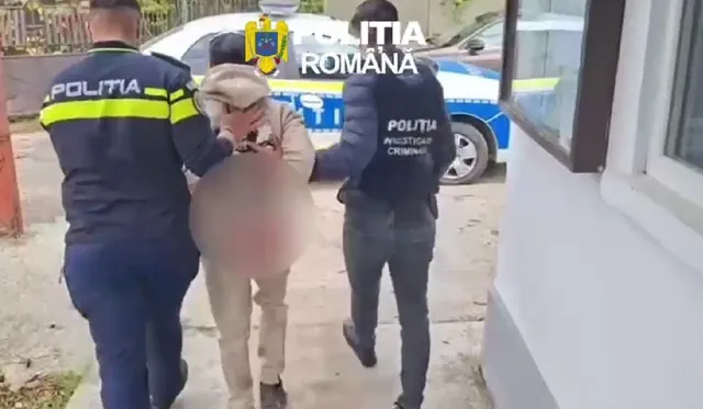 Doi polițiști în uniformă cu inscripția 'POLIȚIA' escortază un bărbat cu fața blurred, îmbrăcat într-o jachetă deschisă la culoare și pantaloni deschiși, lângă o mașină de poliție albastră și albă.