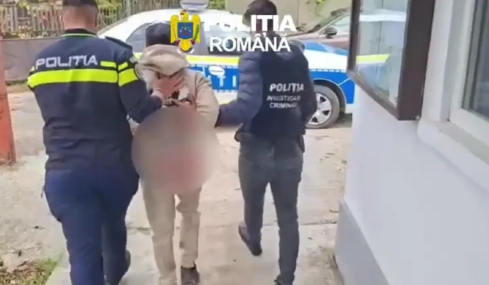 Doi polițiști în uniformă cu inscripția 'POLIȚIA' escortază un bărbat cu fața blurred, îmbrăcat într-o jachetă deschisă la culoare și pantaloni deschiși, lângă o mașină de poliție albastră și albă.