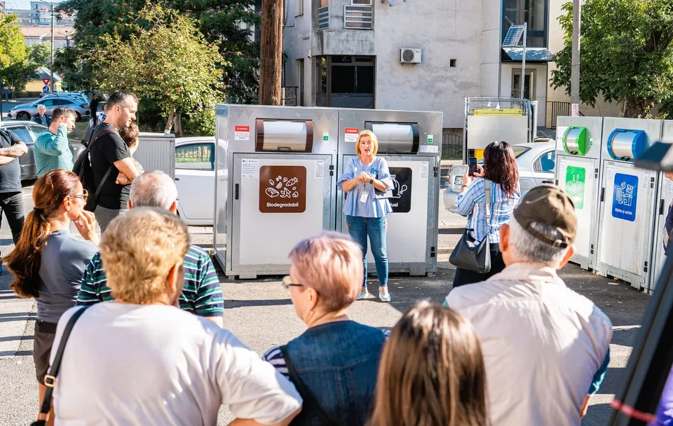 Un grup de persoane stă în fața unor containere metalice de colectare a deșeurilor, în timp ce o femeie stă în fața acestora.
