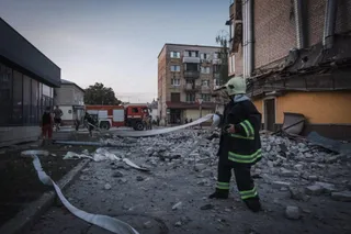 Un salvator cu cască și vestă reflectorizantă merge printre dărâmături în fața unei clădiri avariate sub un cer deschis.