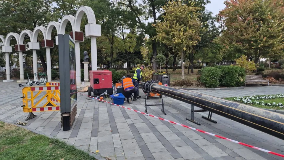 O conductă lungă și neagră este așezată pe suporturi metalice pe un drum pavat, lângă un lucrător purtând vestă portocalie și un echipament roșu.