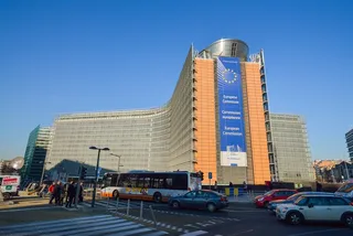 O clădire înaltă cu o fațadă curbată și un banner albastru vertical este situată lângă o stradă cu autobuze și mașini sub un cer senin.