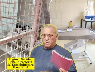 Un bărbat cu ochelari și un dosar roz stă în fața unei mese metalice și a unei cuști cu câini.