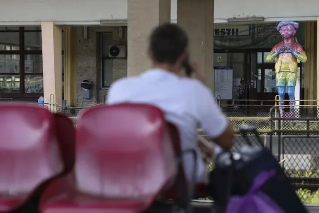 O persoană cu tricou alb stă pe un scaun roșu, în timp ce în fundal se observă o statuie colorată lângă o clădire.