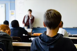 Un grup de elevi stă la bănci în fața unui profesor care se află în fața unei table albe într-o sală de clasă.