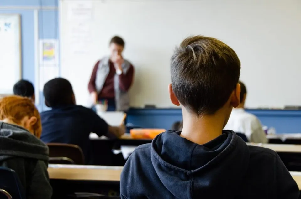 Un grup de elevi stă la bănci în fața unui profesor care se află în fața unei table albe într-o sală de clasă.
