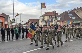 Militari în uniformă de camuflaj defilează pe stradă, purtând steagul României, însoțiți de oficiali și spectatori.