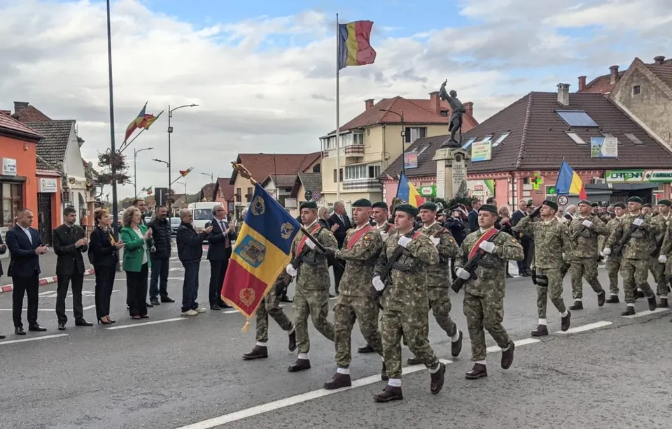 Militari în uniformă de camuflaj defilează pe stradă, purtând steagul României, însoțiți de oficiali și spectatori.