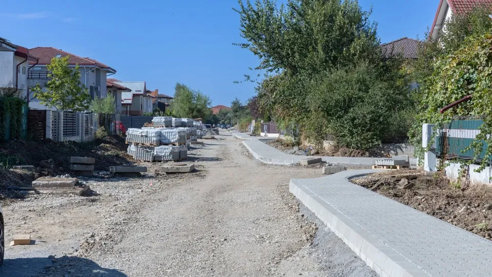 O stradă de pietriș este delimitată de borduri de beton gri și stive de pavaj pe partea stângă, lângă case și vegetație.