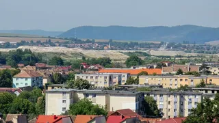 O vedere panoramică asupra unui oraș cu clădiri rezidențiale, case cu acoperișuri colorate și dealuri verzi în fundal sub un cer senin.