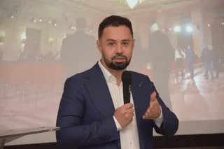 Un bărbat cu barbă, purtând sacou albastru și cămașă albă, ține un microfon negru în mână în fața unui ecran proiectat.