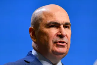Un bărbat cu părul scurt și deschis la tâmple poartă un sacou albastru și o cămașă deschisă, fiind surprins în prim-plan pe un fundal albastru.