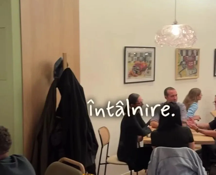 Un grup de persoane stă la o masă într-o încăpere cu tablouri pe perete și un corp de iluminat suspendat.