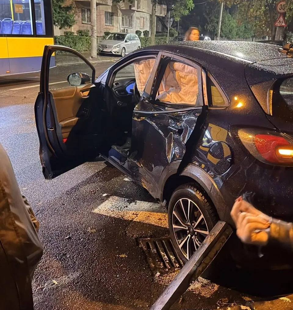 Un autoturism negru cu portierele deschise și airbag-uri gonflate prezintă daune structurale pe partea laterală pe un drum umed noaptea.