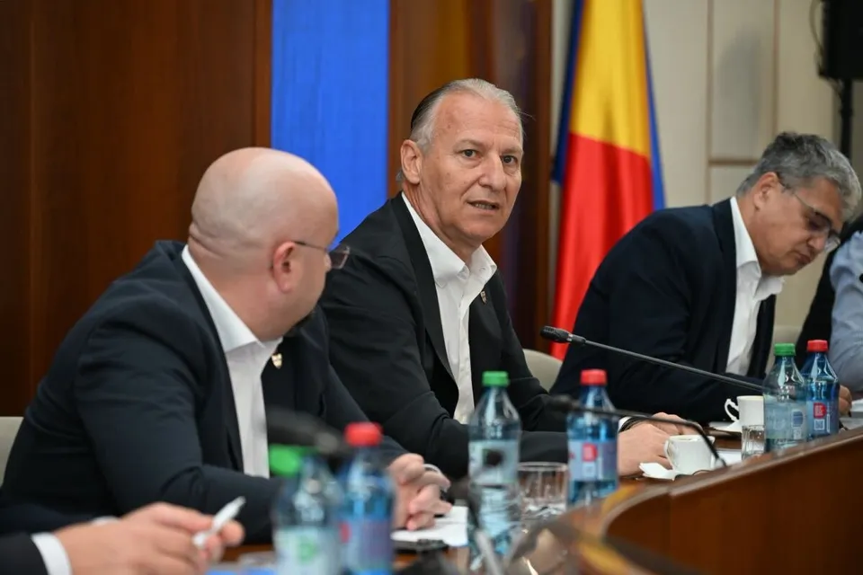 Trei bărbați în costume purtând cămași albe stau la o masă de conferințe cu sticle de apă și microfoane în față.