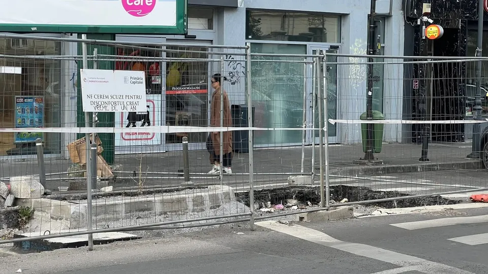 O persoană stă în picioare lângă un gard metalic de șantier din fața unor vitrine de magazine pe un trotuar.
