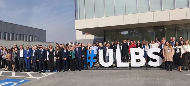 Un grup numeros de persoane stă în fața unei clădiri moderne lângă litere mari albe care formează textul #ULBS.