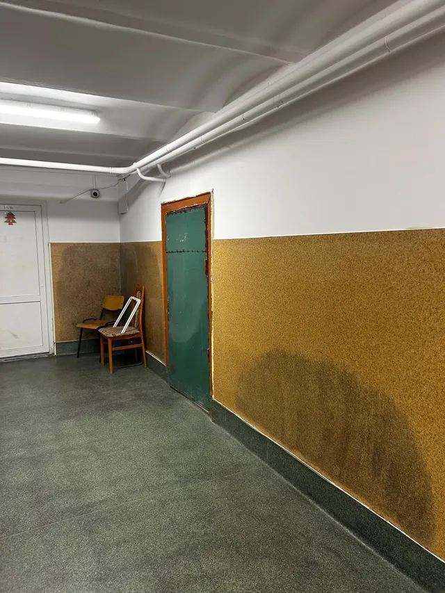 Un hol cu pereți albi și panouri galbene, un podea gri, o ușă verde și două scaune de lemn situate lângă un perete.