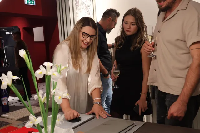 O femeie cu ochelari și bluză albă privește un catalog pe o masă, în timp ce alți oameni stau în apropiere cu pahare în mână.