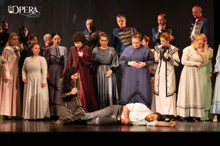 Un grup de actori în costume de epocă stă pe o scenă întunecată, în timp ce un bărbat este întins pe podea în prim-plan.